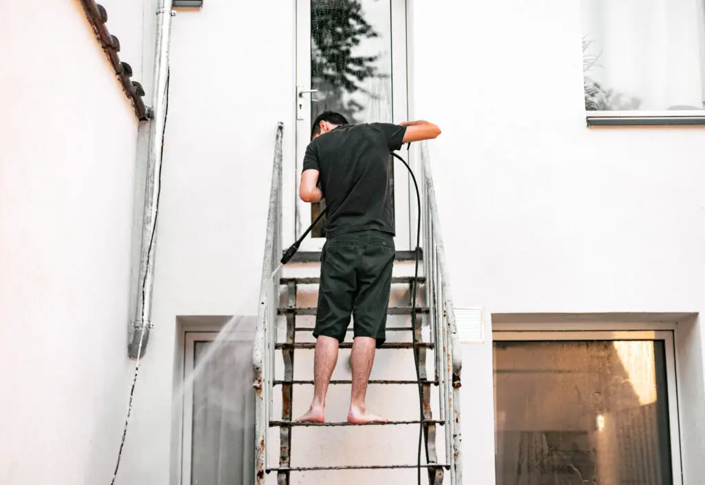 Full length portrait of Caucasian guy standing from behind barefoot in dark clothes washing metal staircase railings with jet of water flying out in splashes under pressure with a kercher in the backyard of his house on a spring day, close-up view from below. Concept of spring start of work using technology.