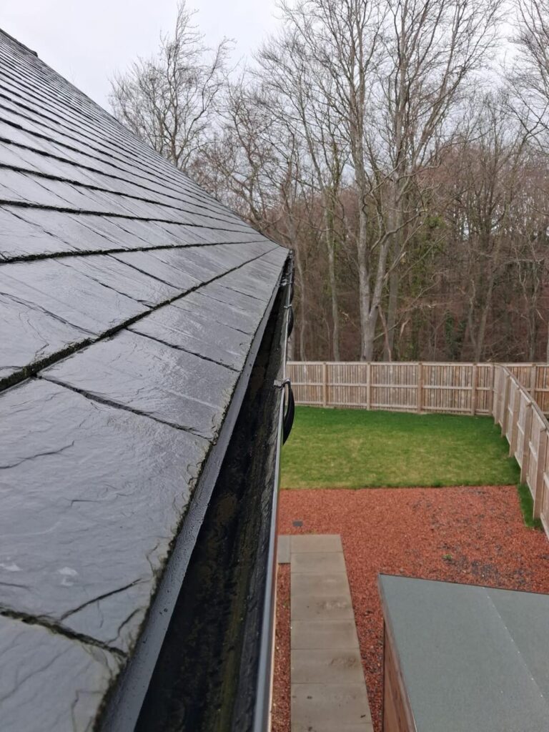 A view from a residential roof featuring a black gutter-cleaning vacuum hose extending along the asphalt shingles, alongside metal mesh gutter guards installed on the rain gutters. This visual represents professional gutter repair and maintenance services.