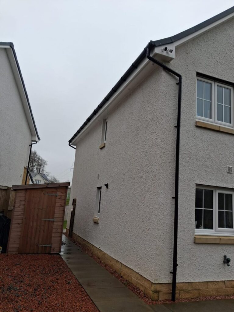 Side view of a white-rendered house with a recently cleaned black drainpipe and gutter system, marked with a blue checkmark to signify completed work.
