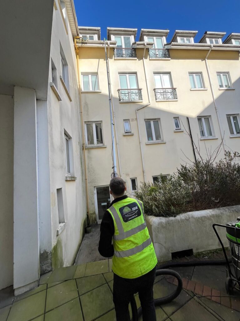 A professional gutter cleaning technician, wearing a safety vest and using a long telescopic pole, works to clear the gutter system of a large, multi-story commercial residential complex.