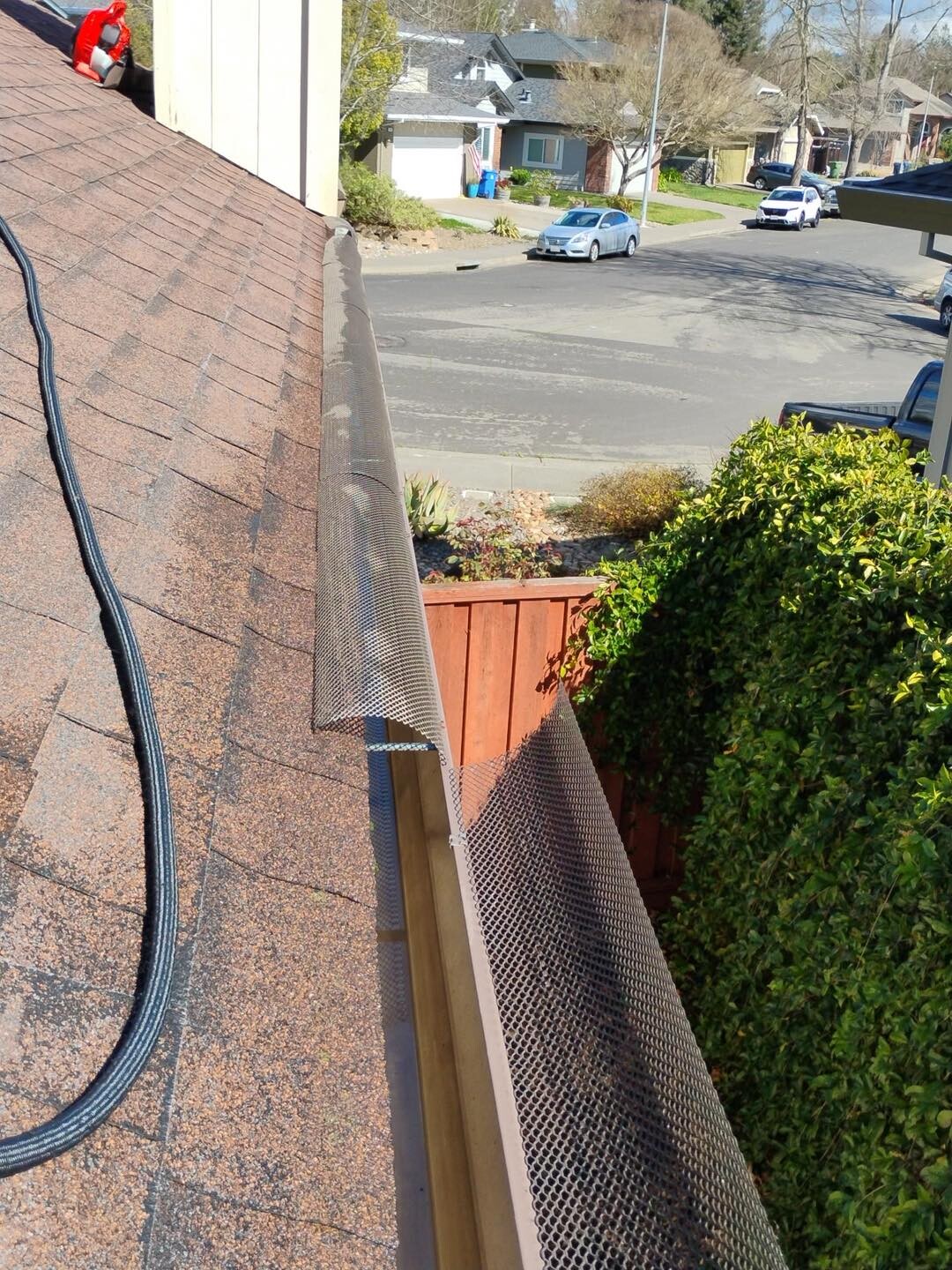 View from a residential asphalt shingle roof featuring a black gutter-cleaning vacuum hose running along the slope toward gutters covered with metal mesh guards, with a suburban neighborhood street visible in the background.