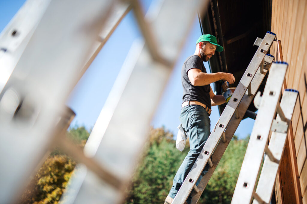 A side-view shot of a professional worker wearing a green cap and work clothes, standing on a ladder and performing repair on a building's roof edge.
