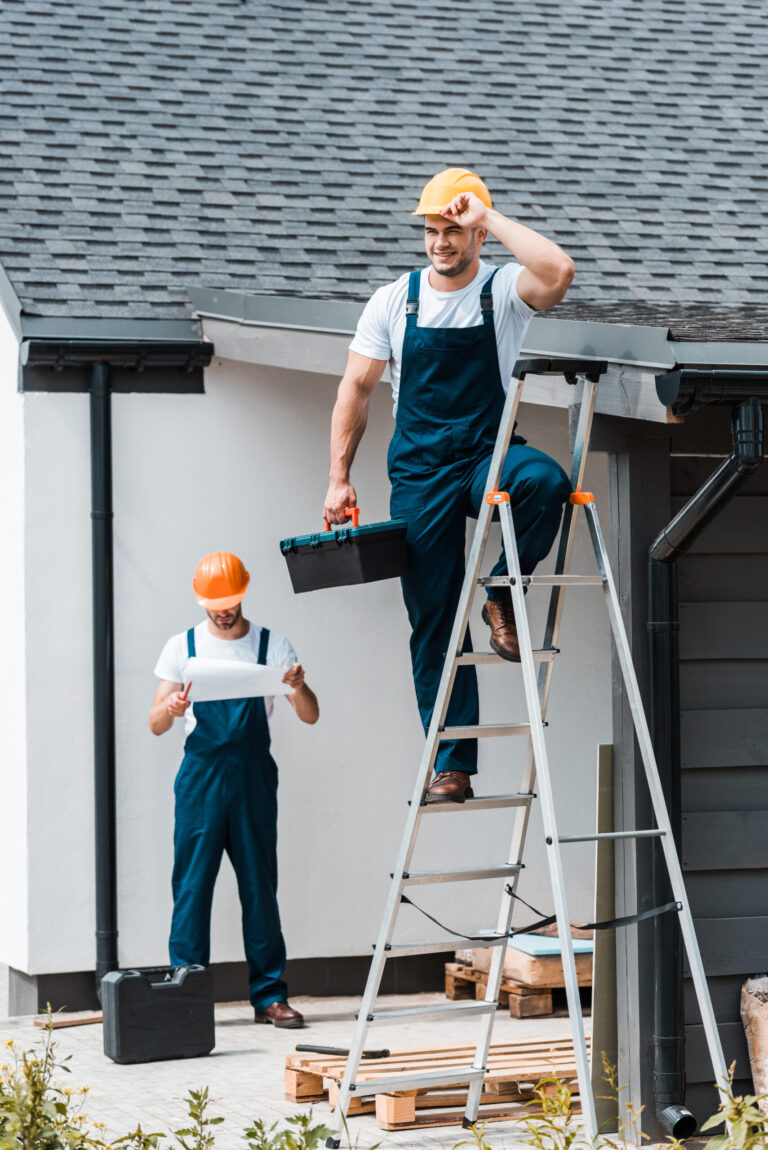 A two-part image displaying a close-up of a residential asphalt shingle roofline on the left, and two professional construction workers in hard hats and blue overalls working on a ladder at the side of a house on the right.
